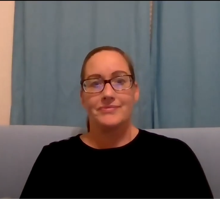A woman in a black shirt wearing glasses is sitting on a blue/gray couch. There are blue curtains behind her with a gold and red pillow to her left.