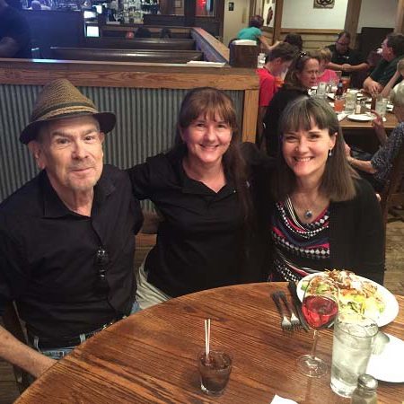 A picture of three of VCD’s Directors from over the years sitting at a table of a restaurant. They are all smiling. Jim Oster, First Director of VCD on the left wearing a hat and a black shirt, Cindy Walsh, current DIrector is in the middle wearing a black shirt ,and Annette Reichman, Director from the late 1990’s is on the right wearing a black, pink, and white shirt. People in the background are eating, with dishes and glasses on the table in front of the group.