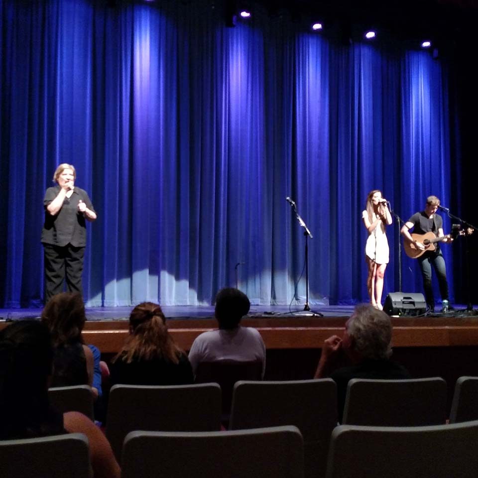 A picture of an auditorium stage. A woman is on the stage to the left, interpreting, in all black. Another woman is in the middle of the stage, singing, with a man playing guitar next to her. There are people in the audience watching.
