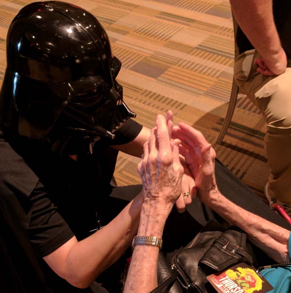 A woman wearing a black mask (Darth Vader from Star Wars) is signing with an elderly woman using tactile sign language.