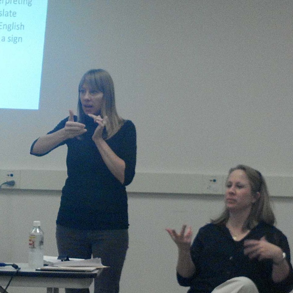 A picture of two women, one is sitting, and one is standing. The woman on the left is standing and interpreting wearing all black clothes. There is a woman sitting to the right wearing all black.