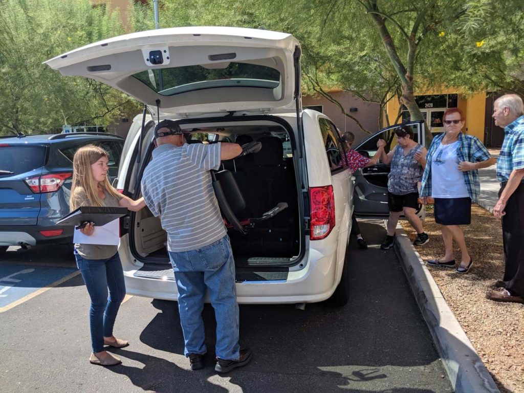 A man and a woman are loading groceries into a van in front of Apache ASL trails. People are getting out of the van and standing to the right of the van. To the left of the van is another parked car.