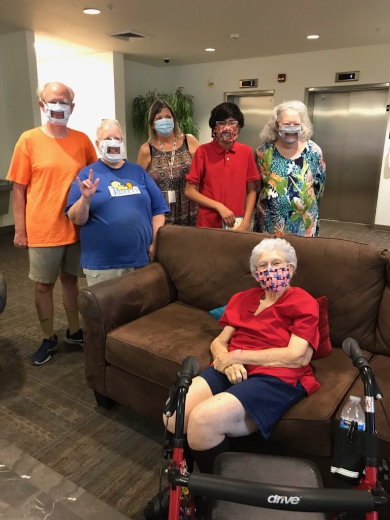 A group of people are standing in the lobby of Apache ASL trails. One woman is sitting on the couch in front of them with her walker in front of her. They are all wearing clear masks.