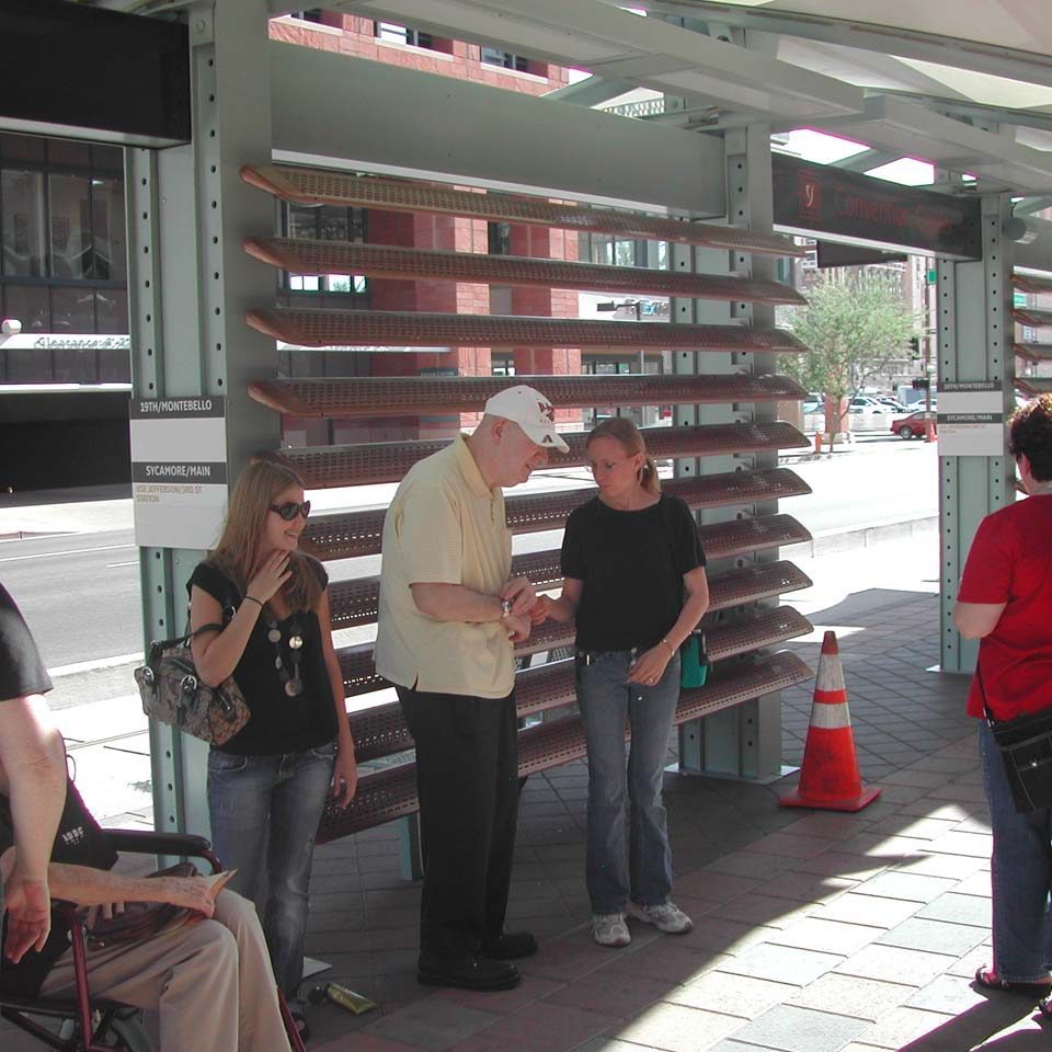 A group of people are standing at the light rail platform. One is guiding a Deafblind individual.