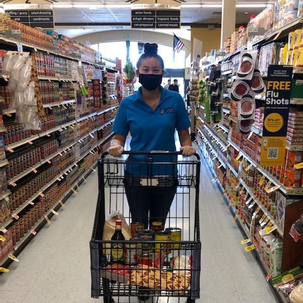 A woman in the supermarket is pushing a shopping cart down the grocery aisle and wearing a blue shirt and a mask.