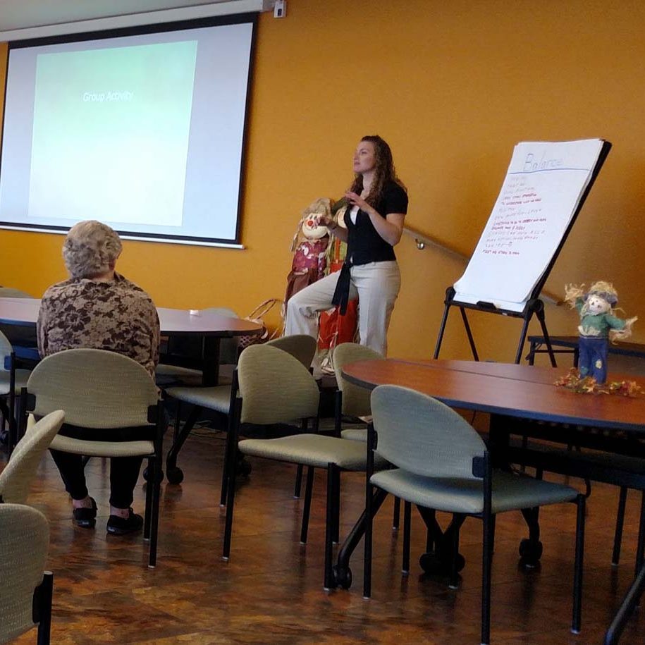 A woman is standing on a stage in the community room at Apache ASL Trails. She is watching a question front the audience. A man is sitting at the table in front of her and a projector screen on the wall behind her. An easel is to the right of her and another table.