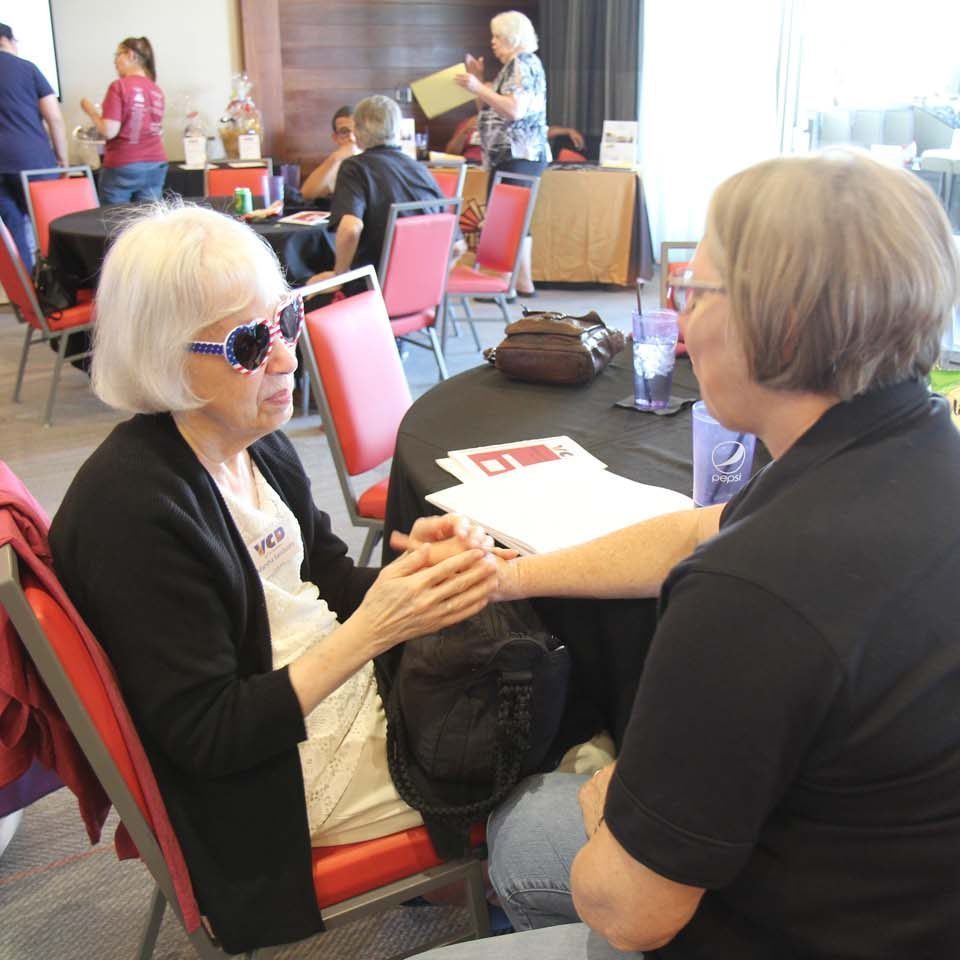 Two women are sitting at a table talking to each other using sign tactile sign language. One is sitting to the left wearing glasses and a white shirt with black jacket and her hands are on top of the interpreter’s hands. The interpreter is wearing a black shirt and glasses. There are people in the background at other tables.