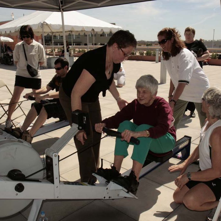 A group of people are on the Ability360 center rooftop. Rowing stations are set up. A woman is standing next to another woman who is sitting at a rowing machine. A woman is behind her and to the right of her assisting her with using the rowing machine. Several people are in the background.